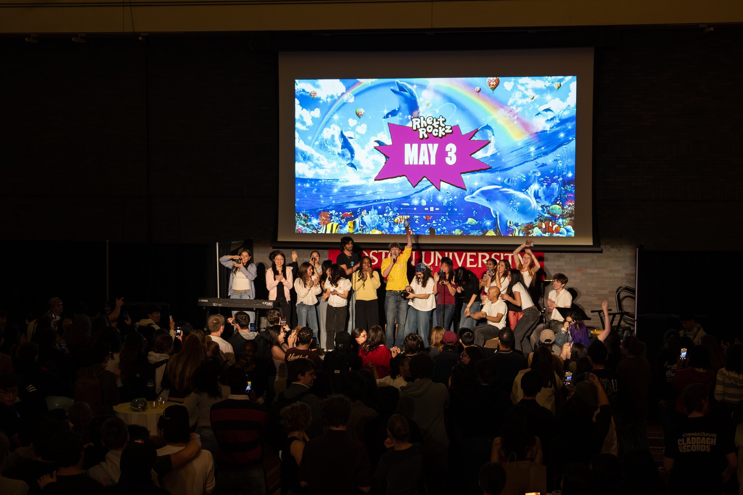 Group of people on stage at an event with a large screen behind them displaying colorful graphics and the words 'Rhett Rockz May 3' in a pink starburst shape.