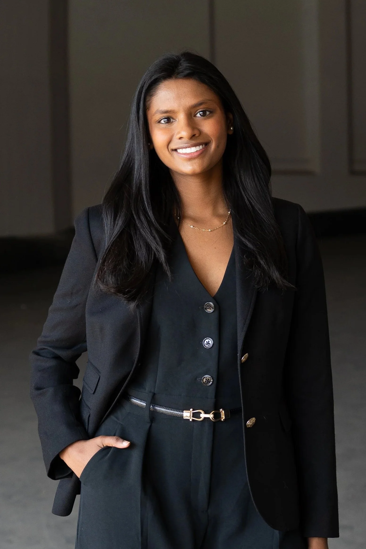 A woman in a black blazer and dress with a gold belt, standing confidently with a smile.