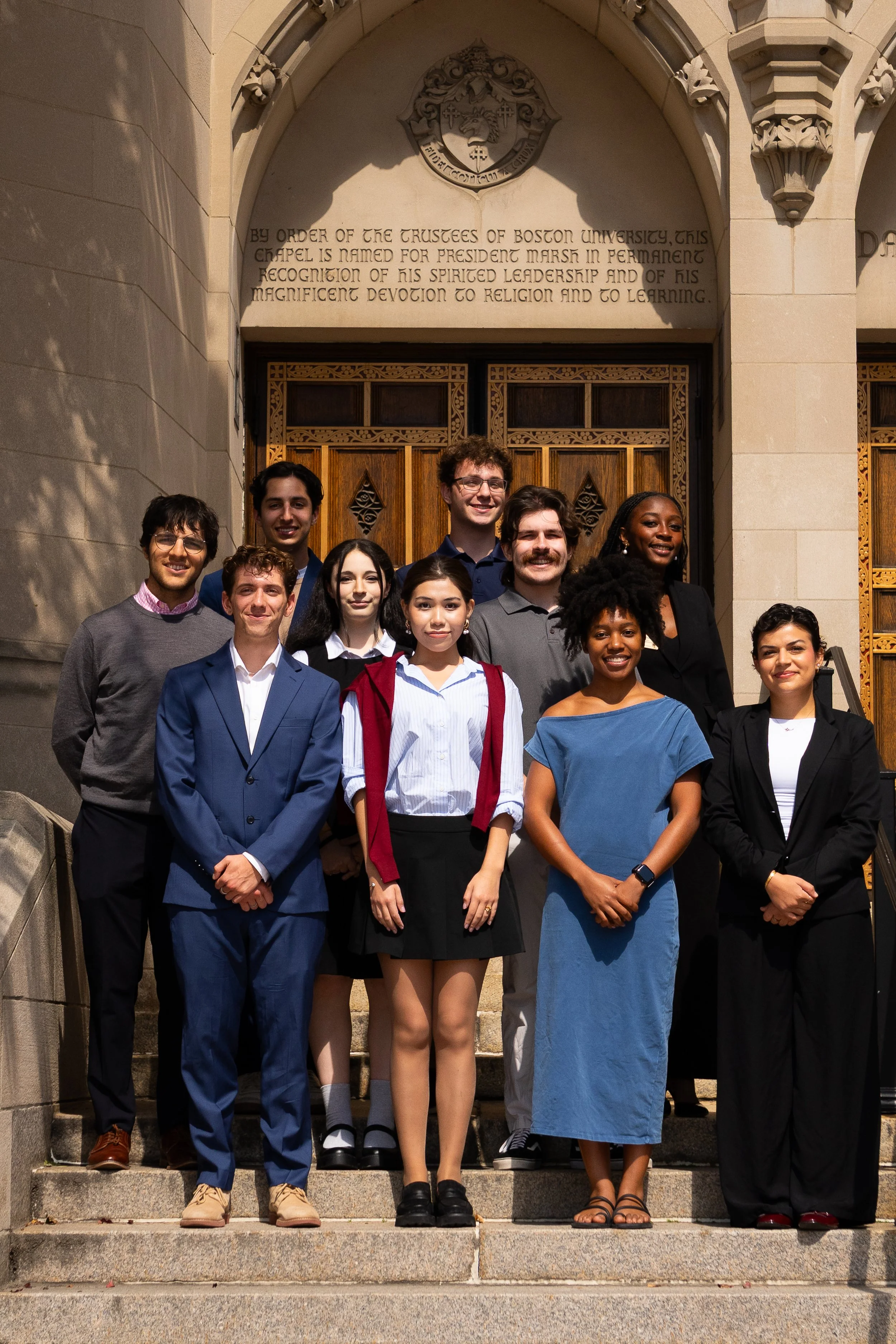 A diverse group of ten young people standing on the steps of a building with stone walls and wooden doors, posing for a group photo. The building has a crest and an inscription above the doorway.