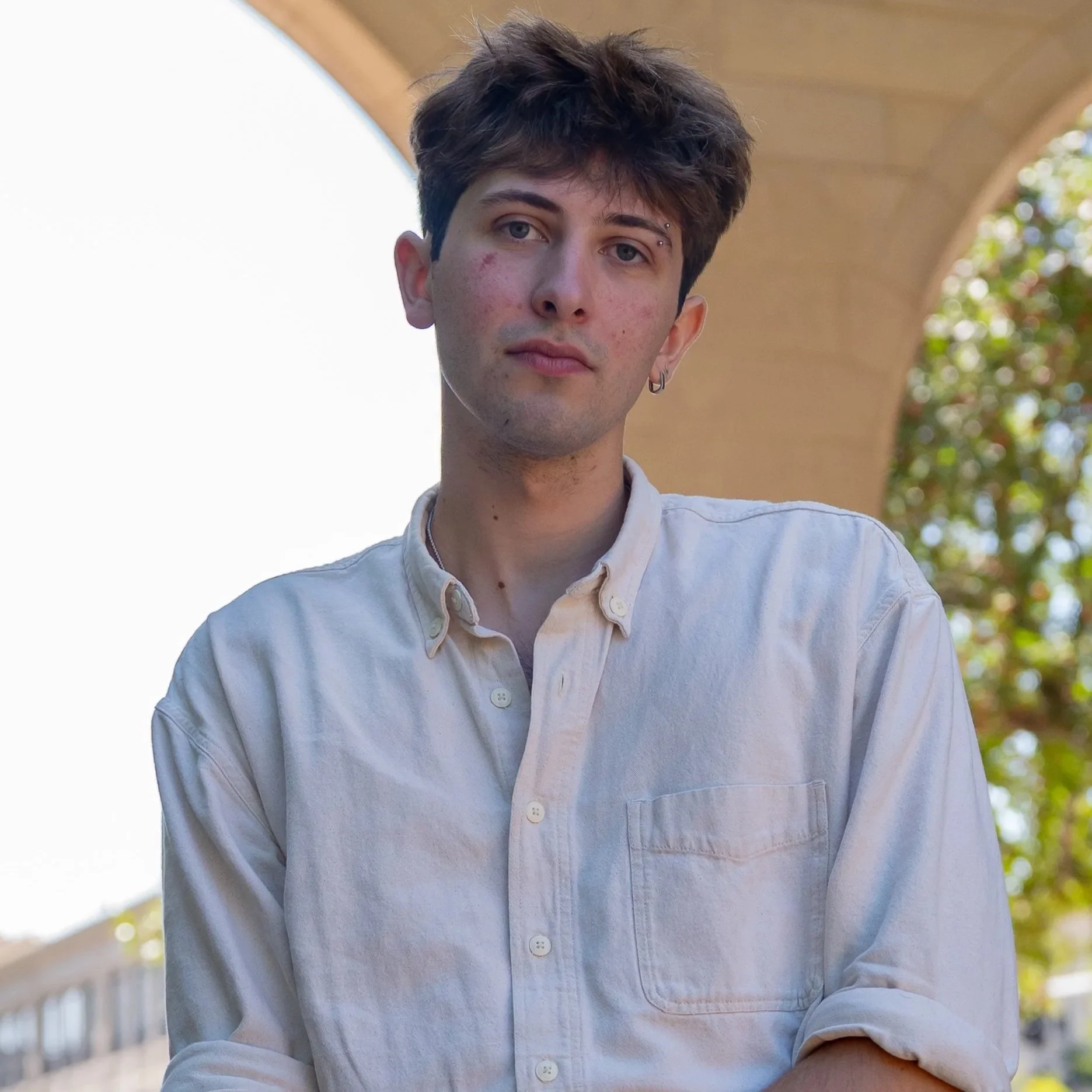 A young man with light skin, brown hair, and light-colored eyes looks directly at the camera. He has some acne on his face, wears a white shirt with a dark sweater over it, and a small earring in his left ear.
