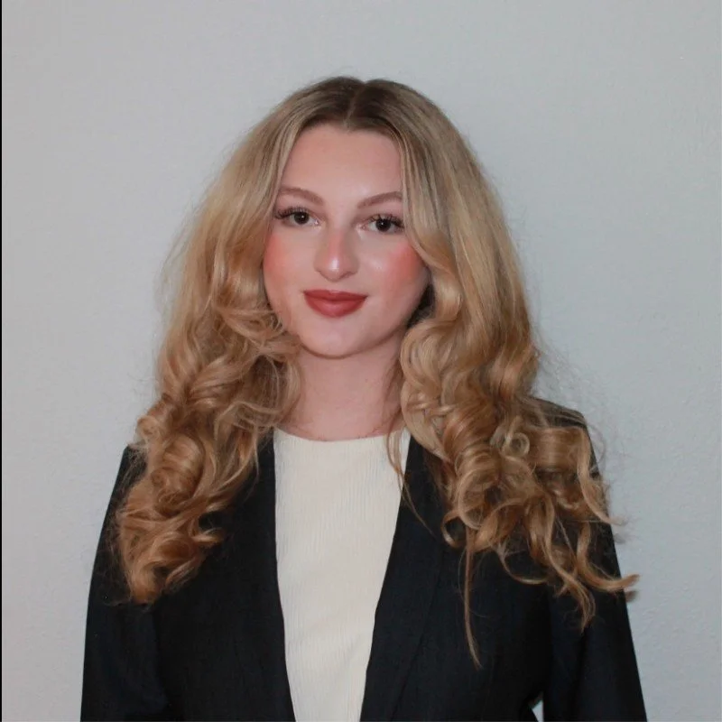 A woman with long, curly blonde hair, wearing a black blazer and a white top, standing against a plain white wall.