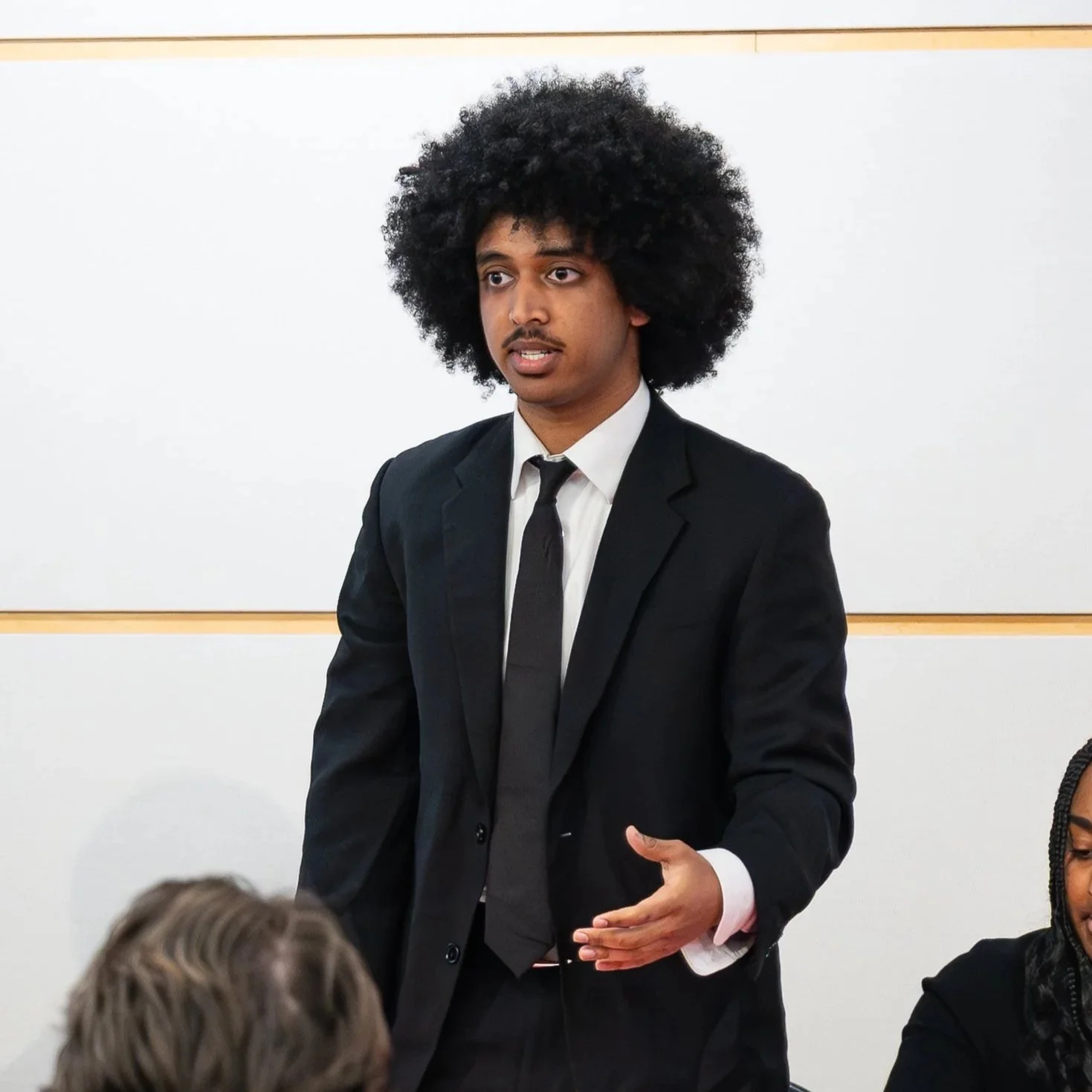 A young man with an Afro hairstyle wearing a black suit, white dress shirt, and black tie, speaking in front of a whiteboard in a classroom or conference room.