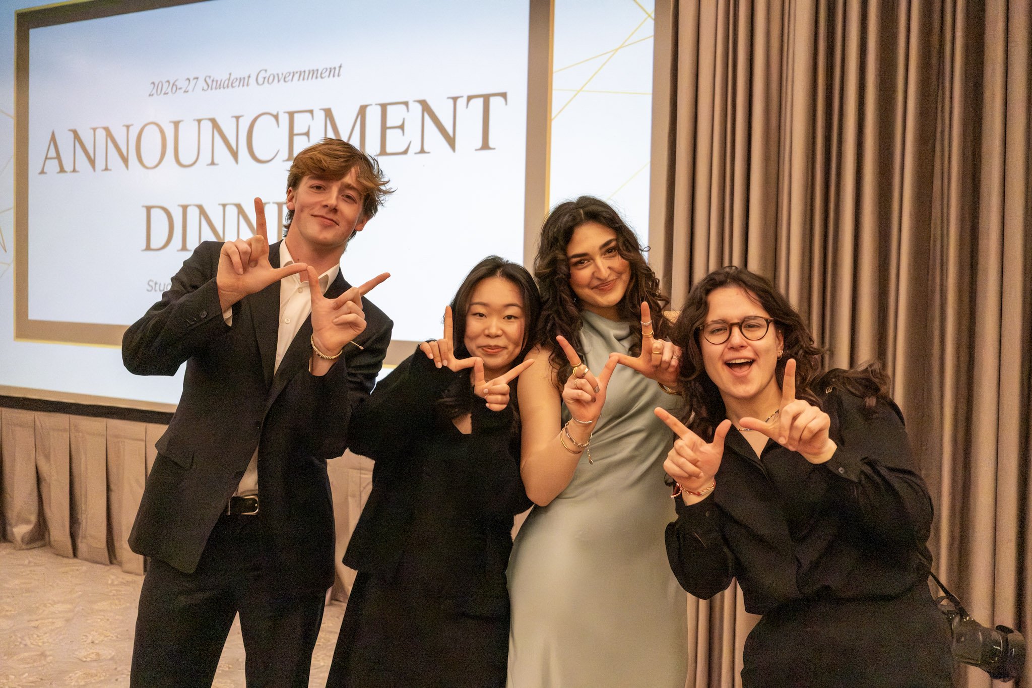 Group of four young adults posing in front of a presentation screen with a slide titled '2026-27 Student Government Announcement Dinner.' They are smiling and making hand gestures.