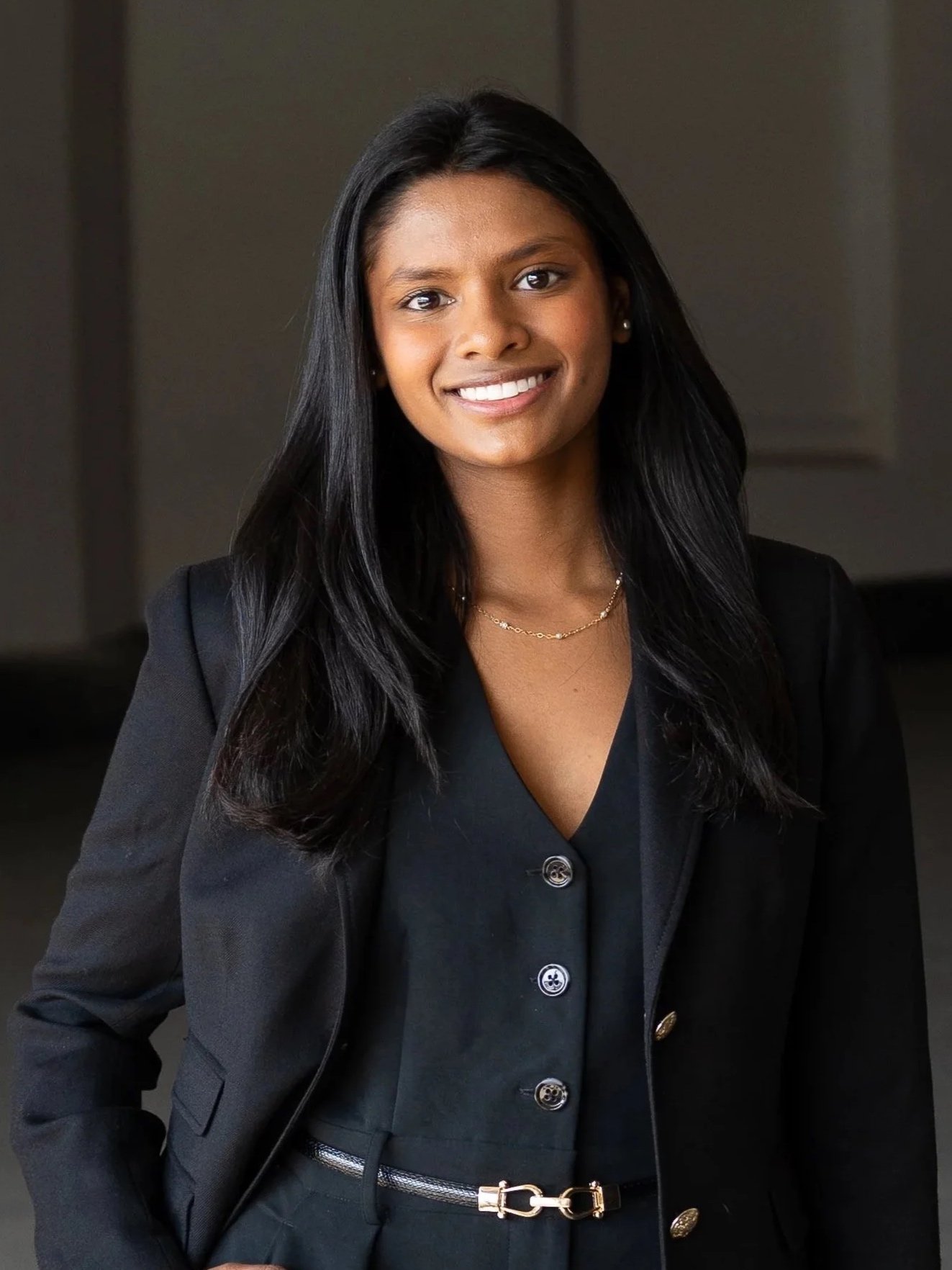 Portrait of a young woman with long black hair wearing a black blazer and a belt with a gold chain buckle, smiling at the camera.