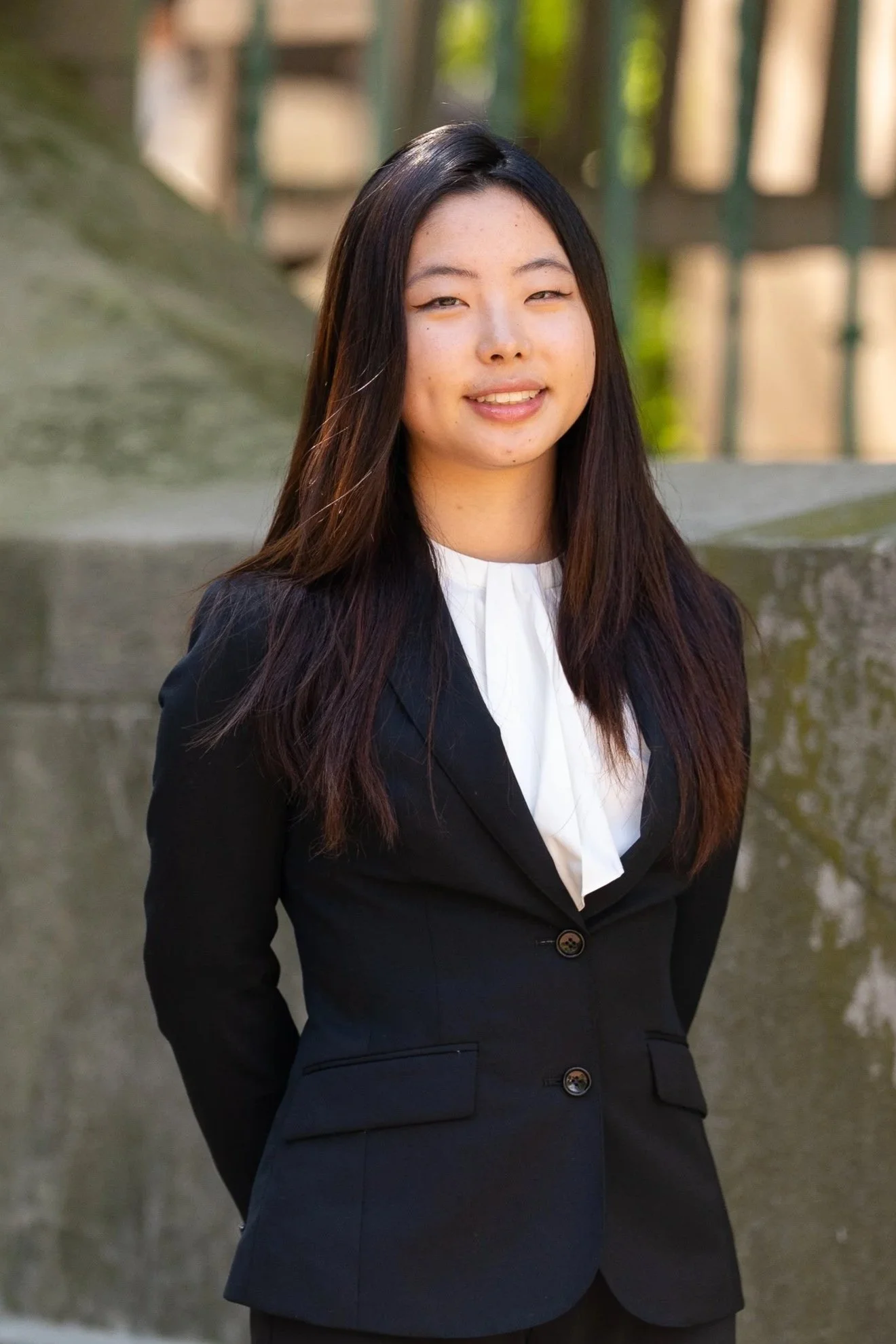 A woman with long dark hair wearing a black blazer and white blouse, standing outdoors near a stone structure and a green metal fence.