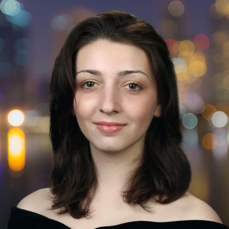 A woman with shoulder-length dark brown hair and fair skin, smiling softly, with a blurred cityscape and colorful lights in the background at night.