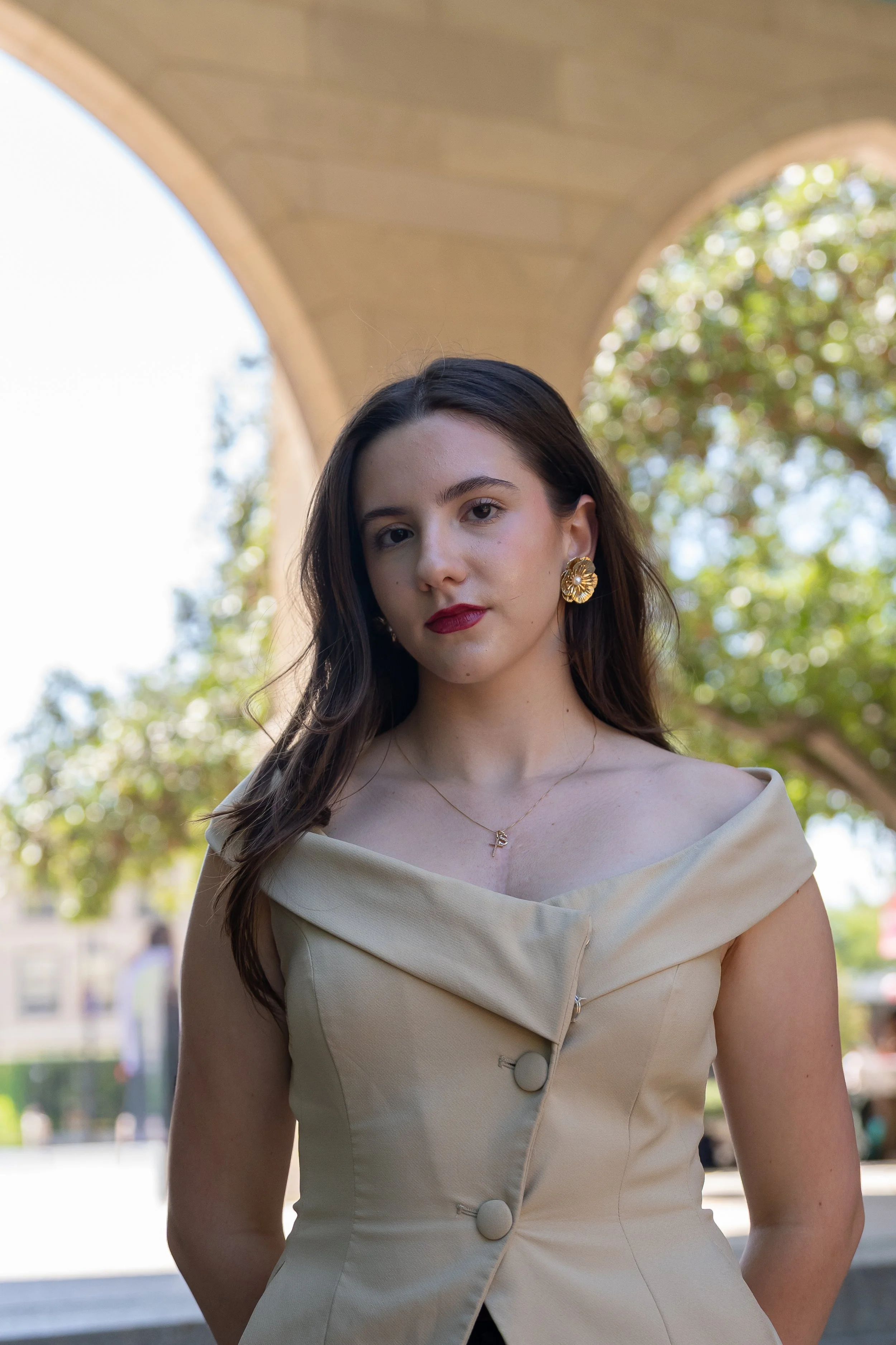 Young woman with long wavy brown hair wearing a light blue blouse, standing against a gray stone wall.