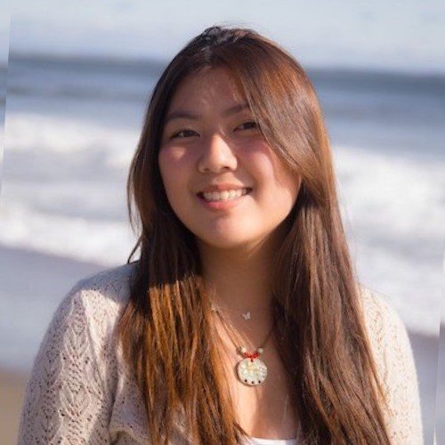Young woman with long brown hair smiling at the beach, wearing a cream sweater and a necklace.