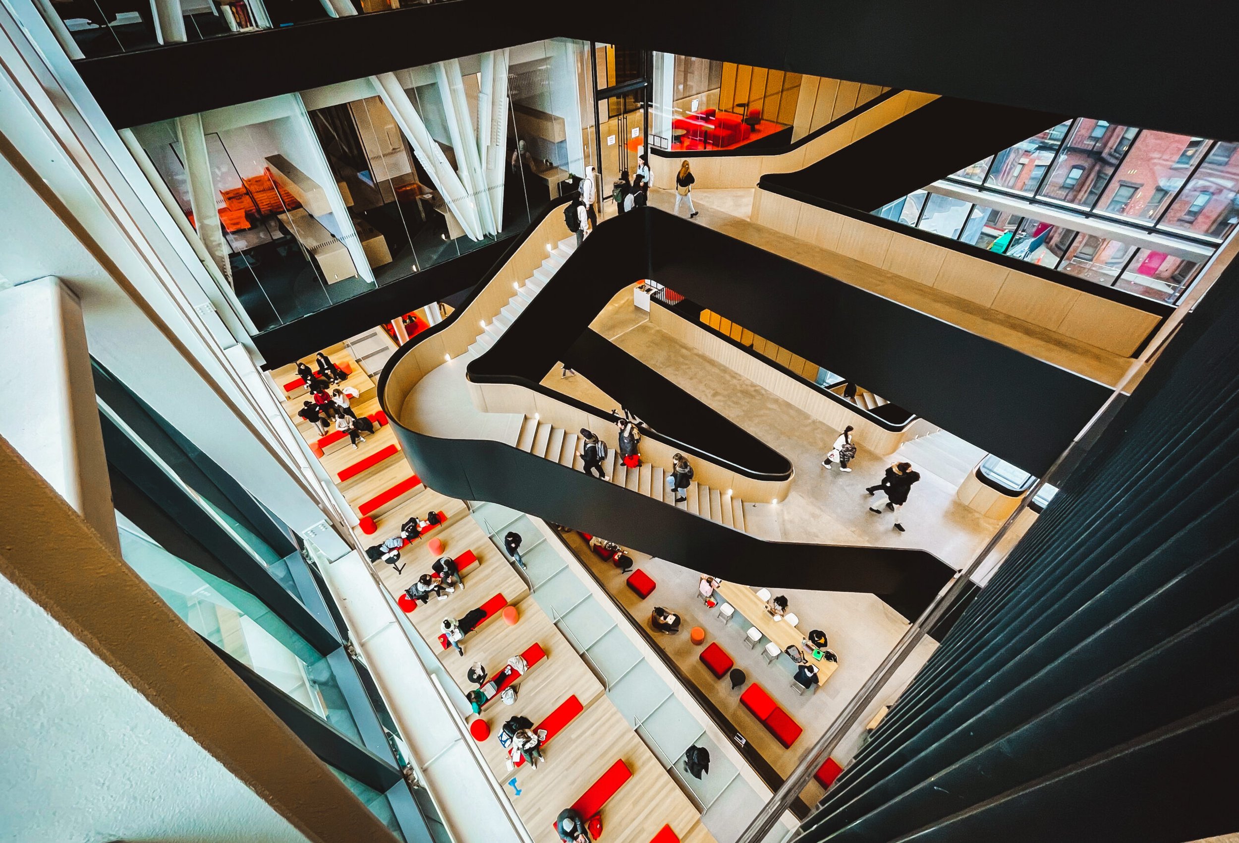 Multiple floors of a modern building with open atrium, people walking, sitting, and talking in the public spaces, with large windows overlooking city streets.