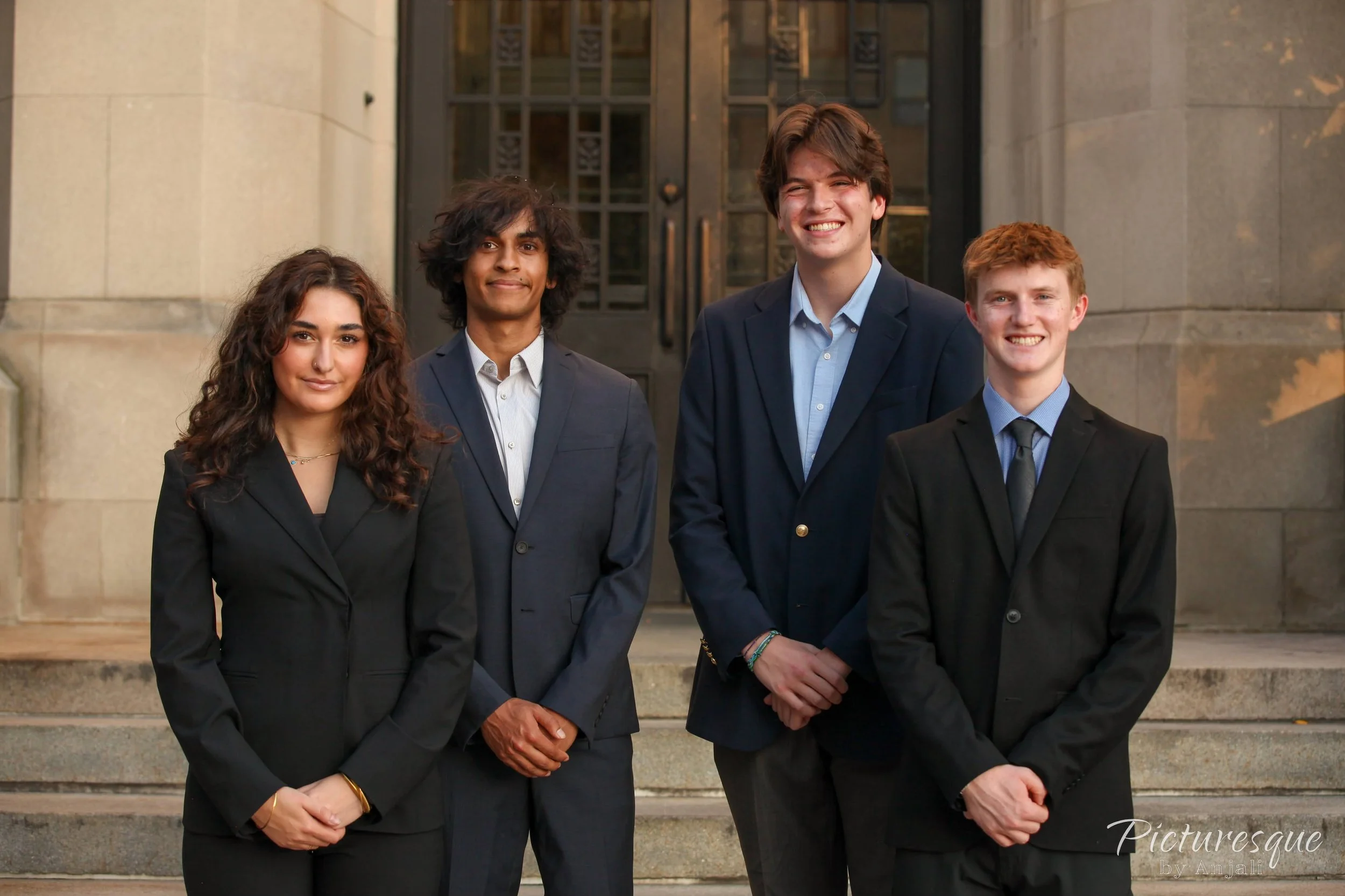 Four young adults, three men and one woman, standing in front of a stone building with large black doors. They are dressed in formal business attire and smiling at the camera.