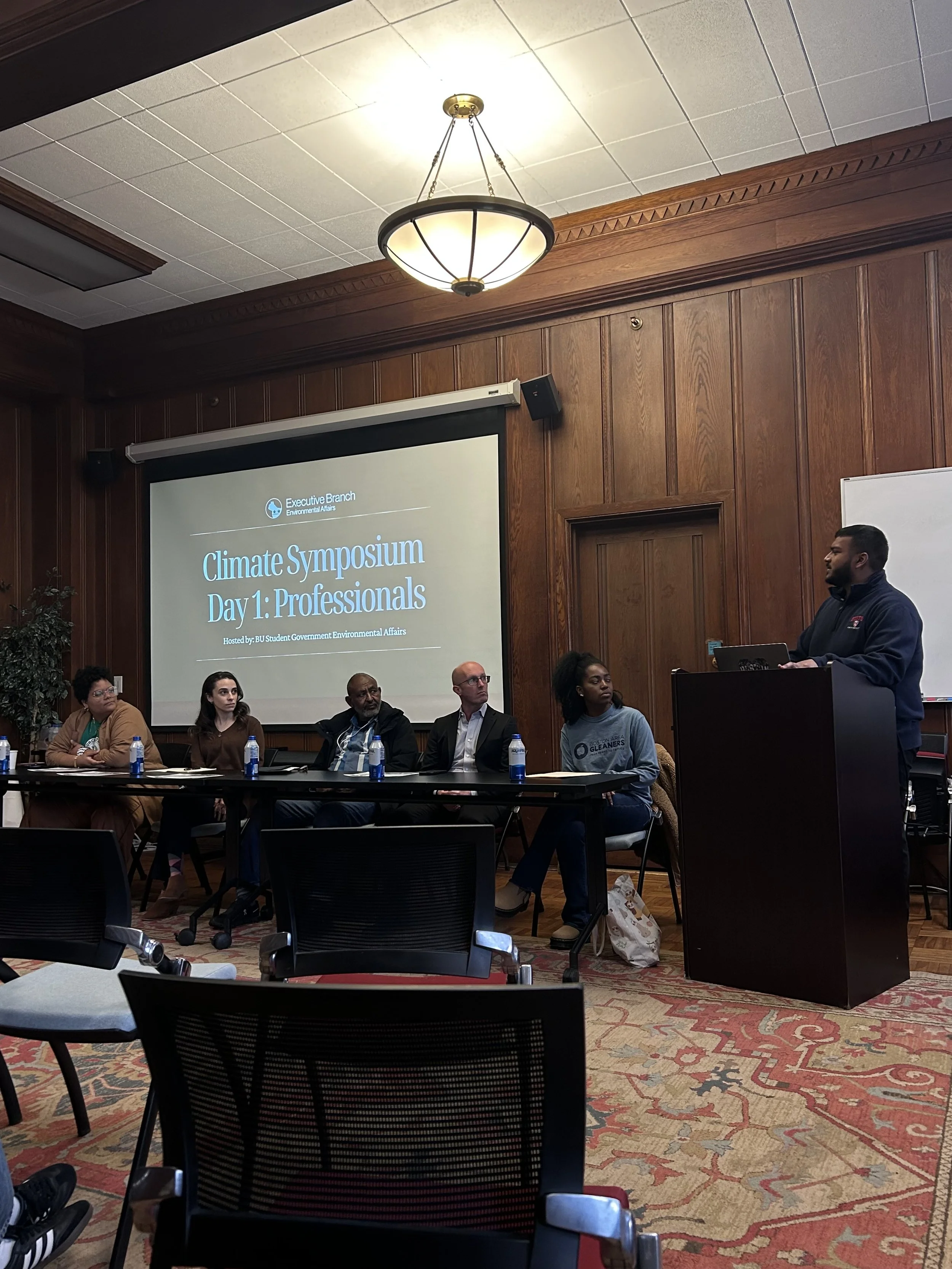 A group of diverse people seated at a long table in a conference room, facing a presentation screen displaying 'Climate Symposium Day 1: Professionals.' A man stands at a podium to the right, speaking. The room has wood-paneled walls, a decorative ceiling, and a large chandelier. Bottled water is placed at the table.