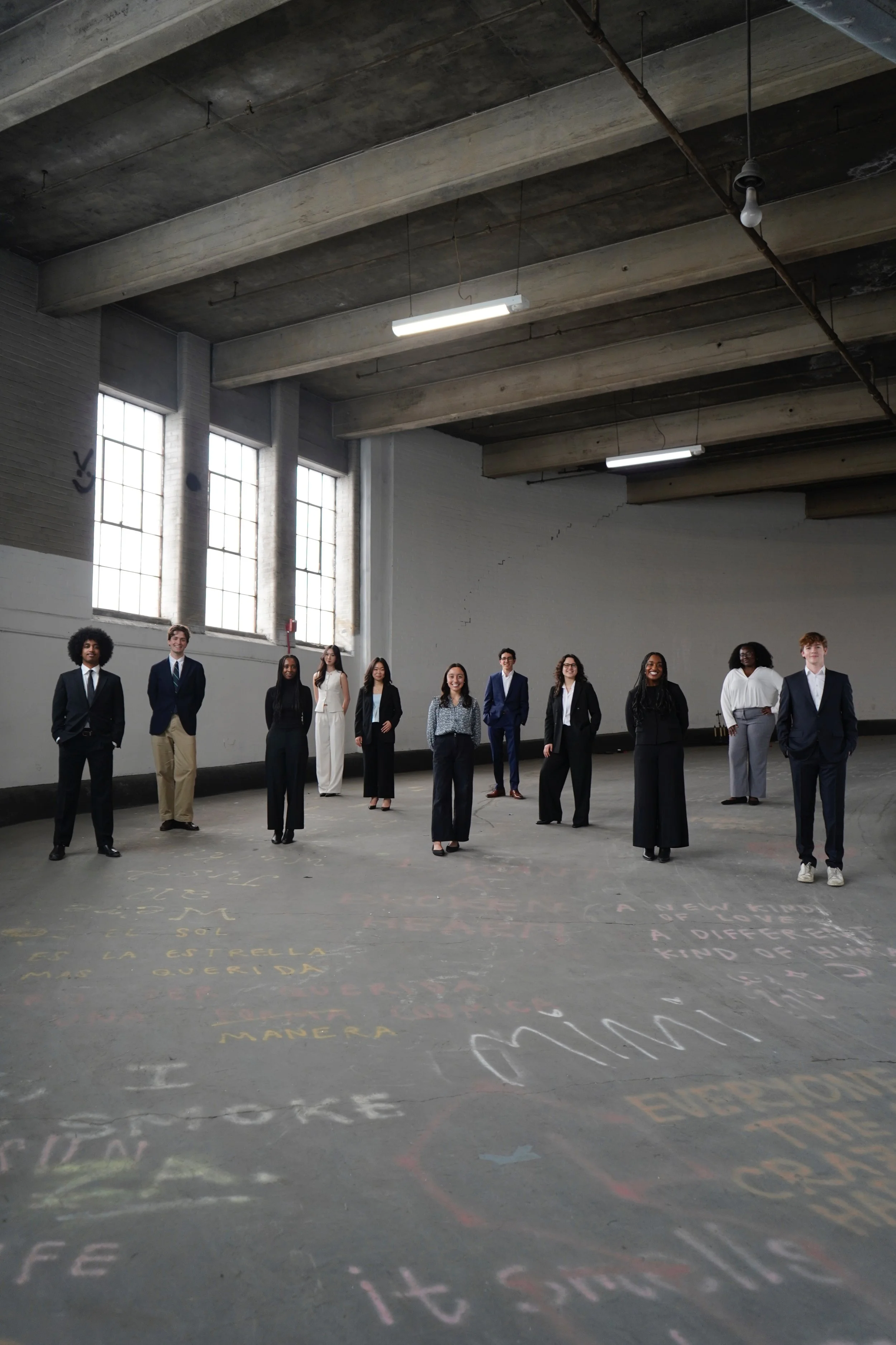 A diverse group of people standing in a spacious, industrial-style room with concrete beams and large windows, smiling at the camera, with colorful chalk writings on the floor.
