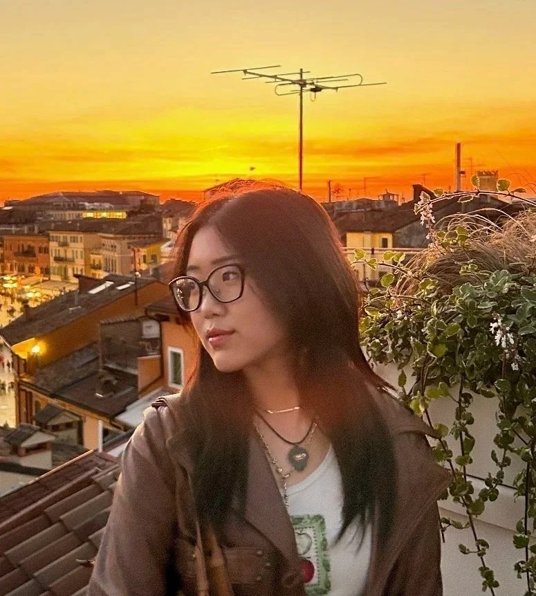 Young woman with glasses and layered necklaces on a balcony during sunset, with city rooftops and a TV antenna in the background.