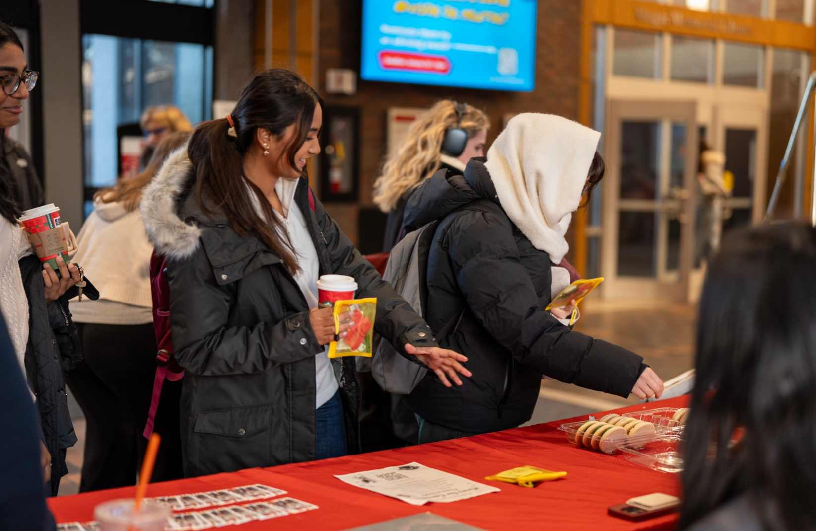 People in line at a food or snack stand inside a building, with some holding coffee cups and browsing items on a table covered with a red tablecloth.