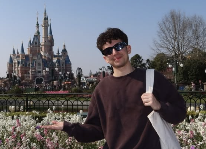 Young man with curly hair and sunglasses standing outdoors in front of Sleeping Beauty Castle at Disneyland, holding out his hand towards colorful flower beds.