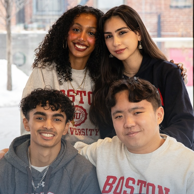 Four diverse young adults smiling and posing together indoors, with a snowy window view in the background.