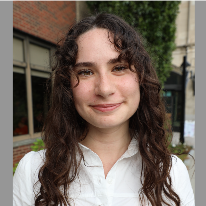 A young woman with curly brown hair and a white button-up shirt, smiling outdoors with a background of a building and trees.