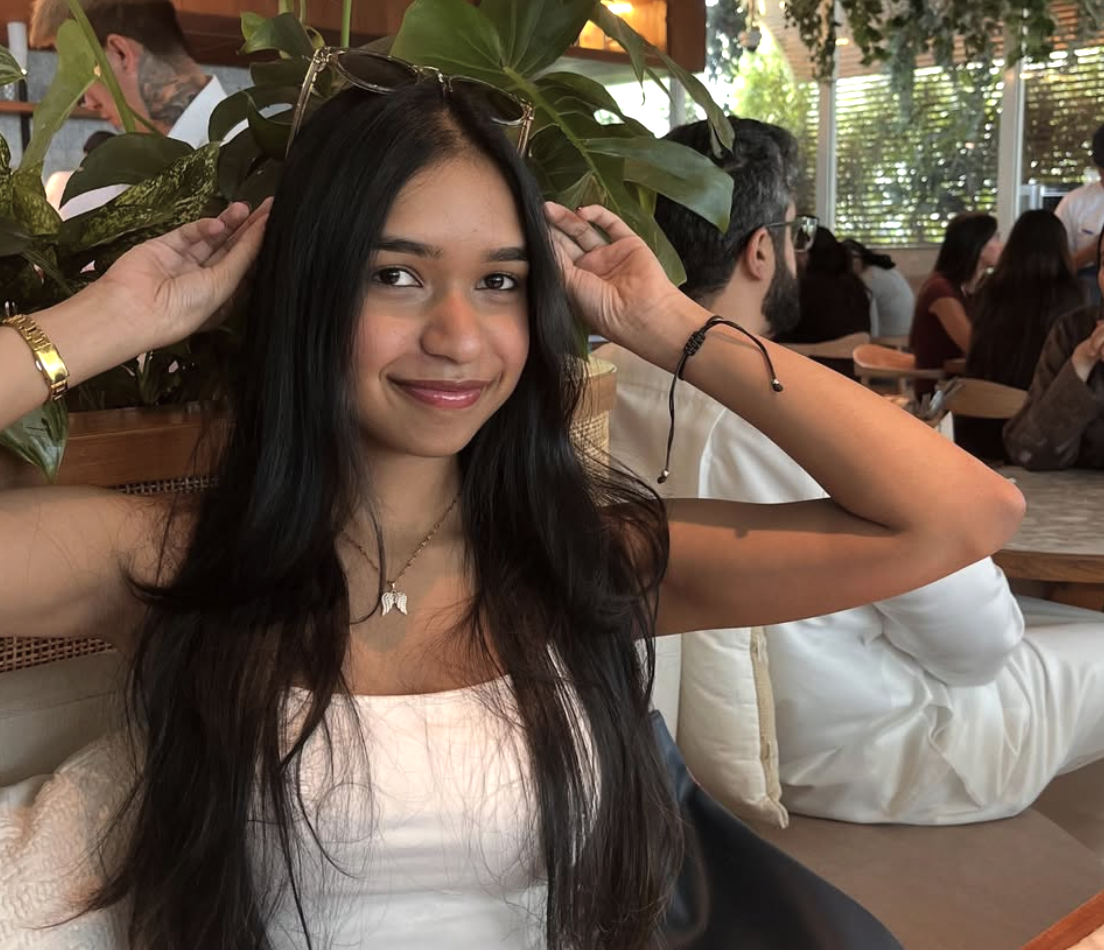 A young woman with long dark hair smiling, sitting in a lively cafe or restaurant with large leafy plants and other people dining in the background.