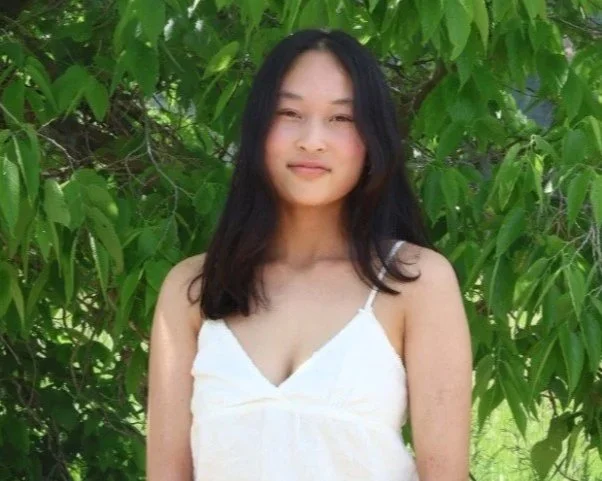 Young woman with long dark hair wearing a white spaghetti strap dress standing outdoors in front of green foliage.