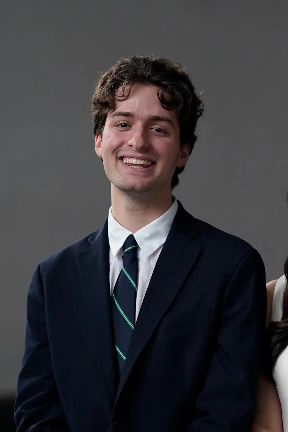 Young man with curly brown hair smiling, wearing a black suit, white shirt, and striped tie, standing against a plain grey background.