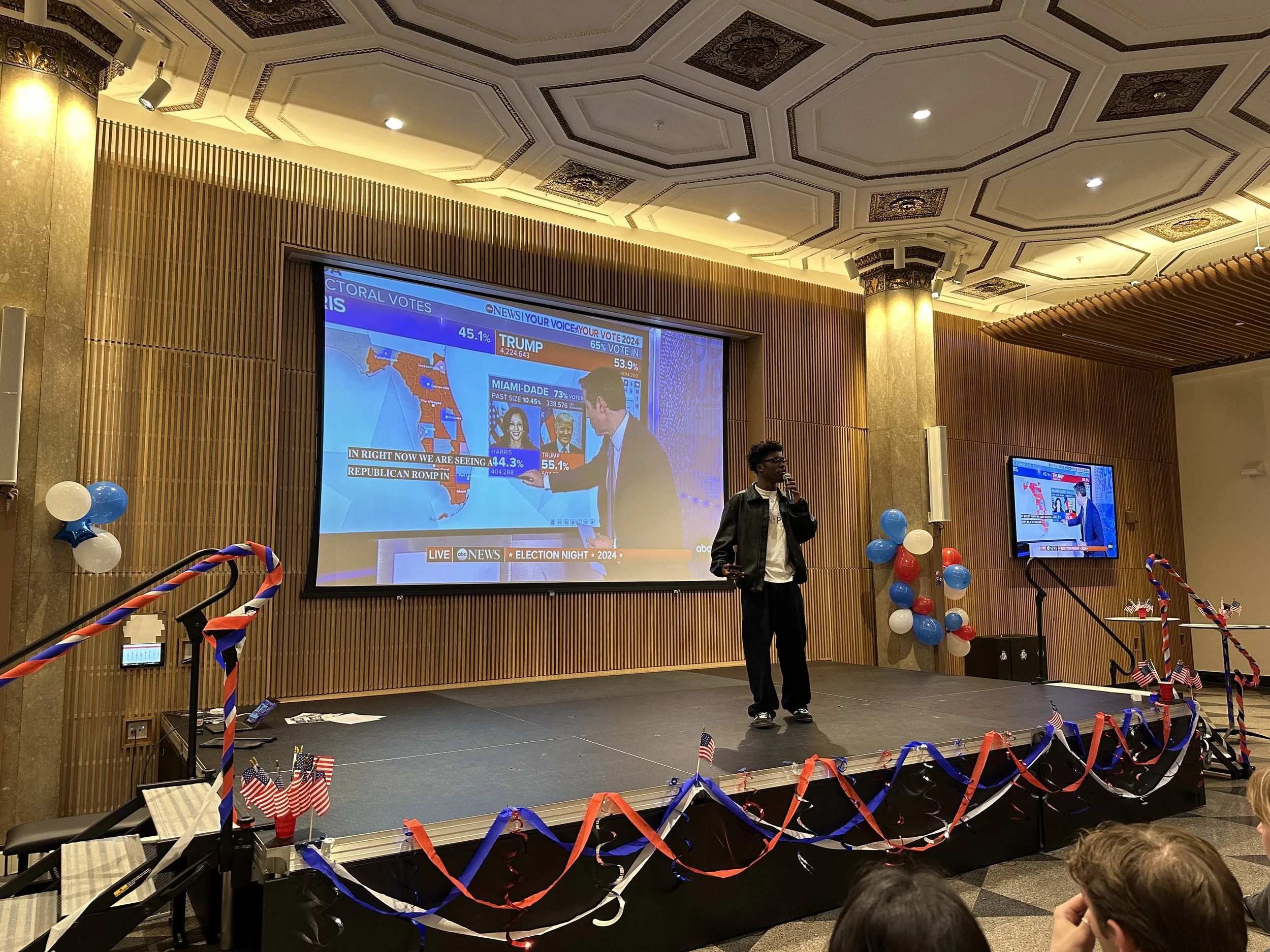 An indoor event with a stage decorated with red, white, and blue balloons and ribbons, American flags, and a large screen displaying election results showing votes for Trump and Harris. A man is speaking into a microphone on stage, and several people are sitting in the audience.