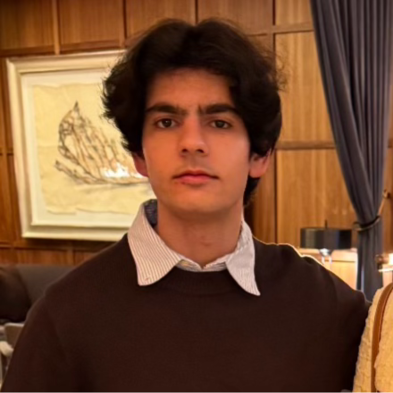 Portrait of a young man with dark, wavy hair, wearing a white collared shirt under a dark sweater, in a room with wooden paneling and artwork on the wall.