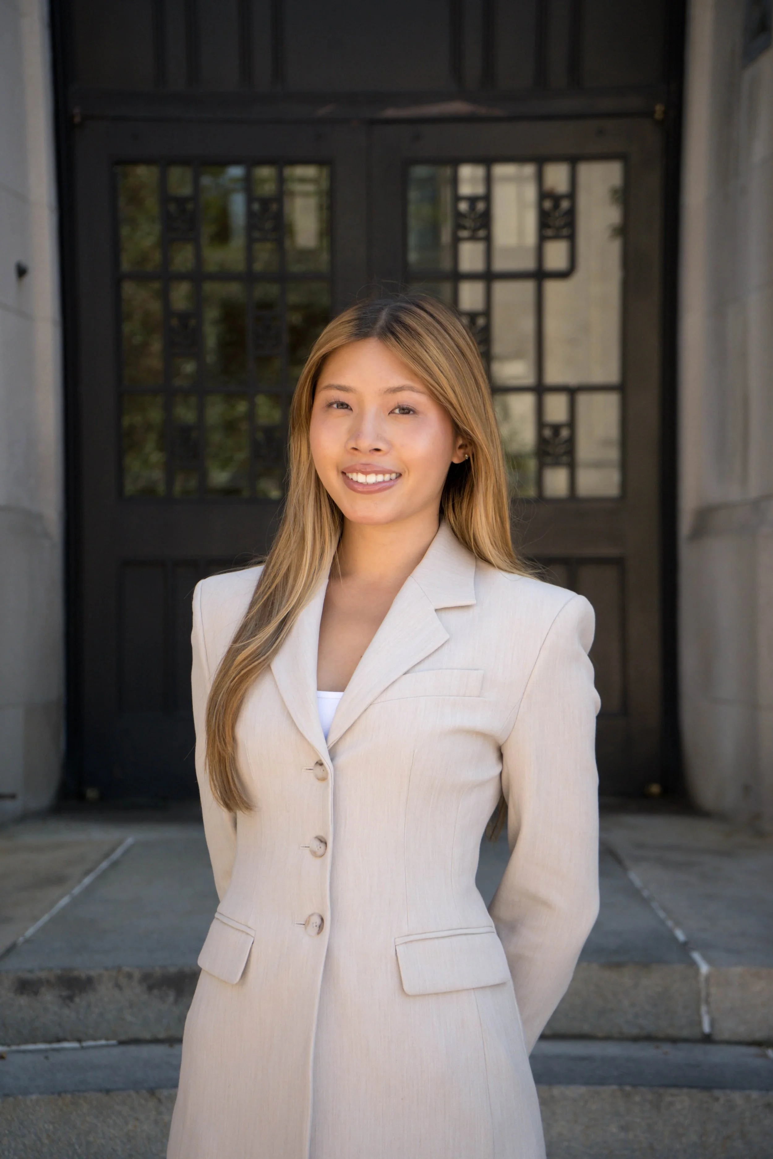 Young woman outdoors with green foliage, smiling and winking, wearing a light-colored blazer and small earrings.