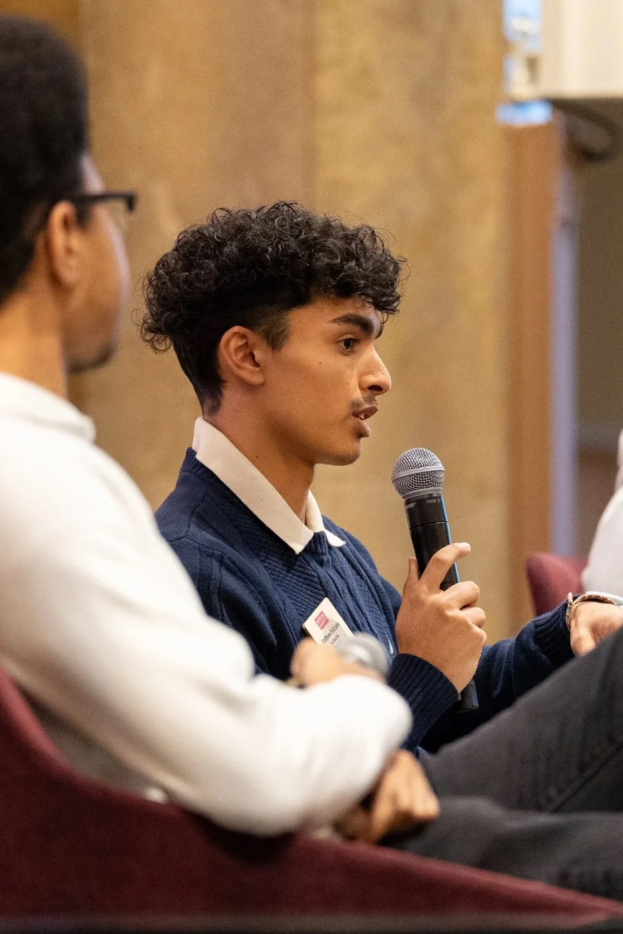 A young man with curly hair and a navy sweater speaking into a microphone during a discussion panel; other participants are visible in the foreground and background.