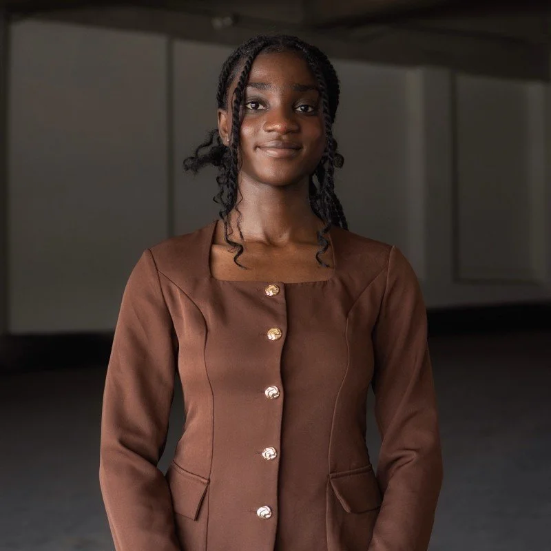 A young African American woman with braided hair wearing a brown blazer with gold buttons, standing indoors in front of a neutral background.