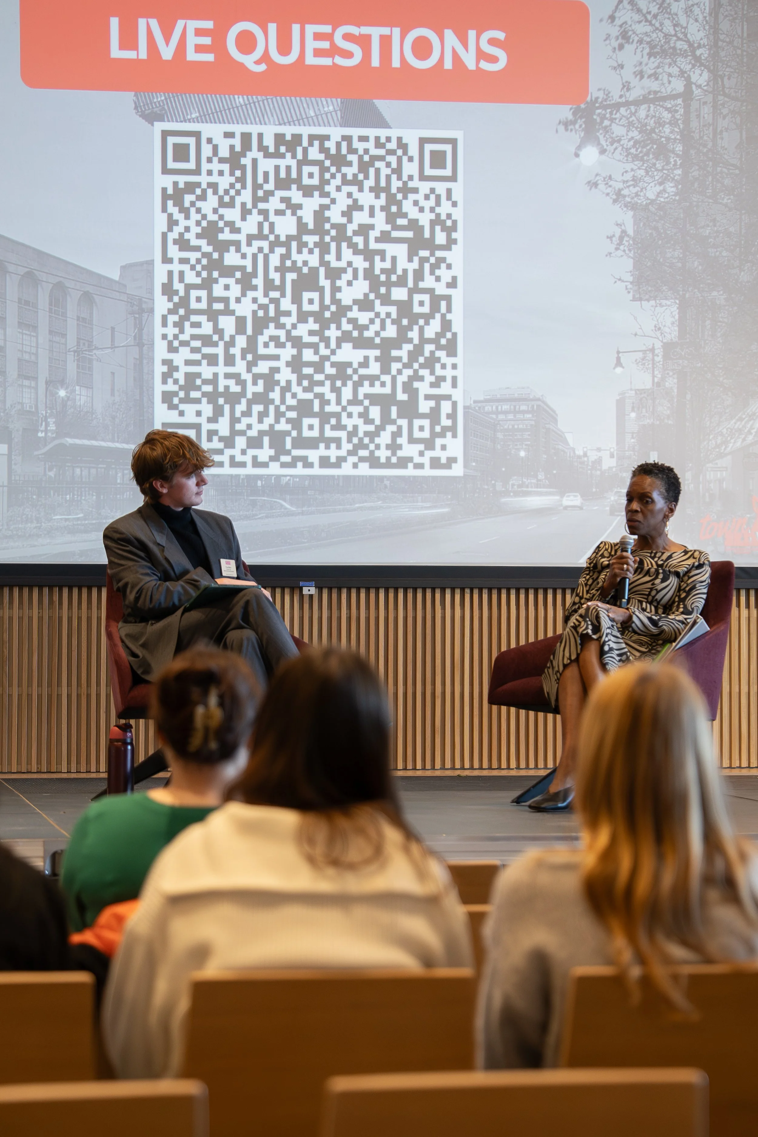 A panel discussion with two women on stage, one speaking into a microphone, in front of an audience. A large screen behind them displays a QR code and the text 'LIVE QUESTIONS' at the top, with a city street background.