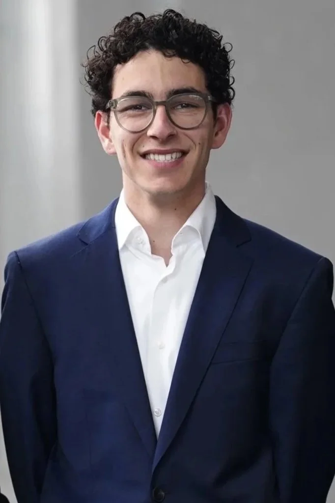 Young man in a navy blue suit and white shirt, smiling and wearing glasses, standing against a plain background.