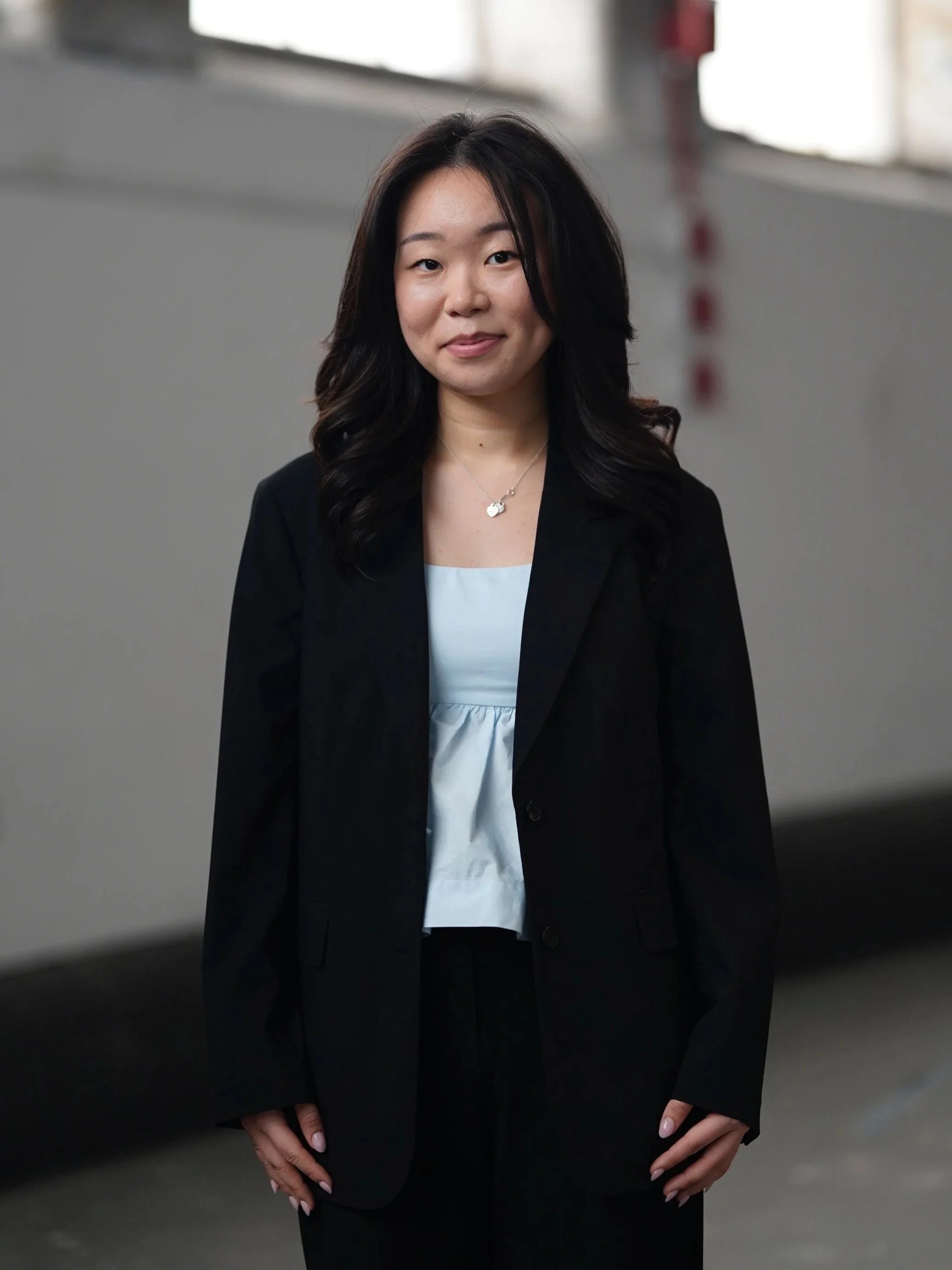 Portrait of a woman with medium-length dark hair, wearing a black blazer with a white top underneath, standing in front of a blurred gray background.