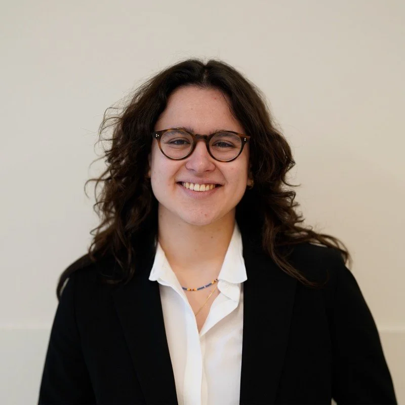 A woman with wavy brown hair, glasses, and a white shirt with a black blazer smiling at the camera.