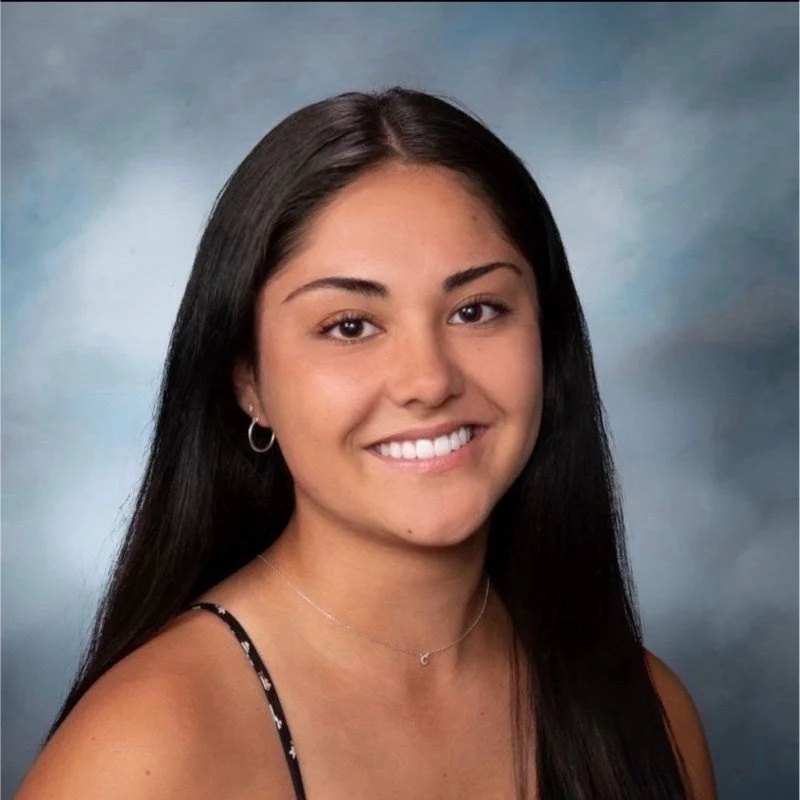 Young woman with long dark hair, wearing a black spaghetti strap top, smiling with white teeth, in front of a blue-gray painted background.