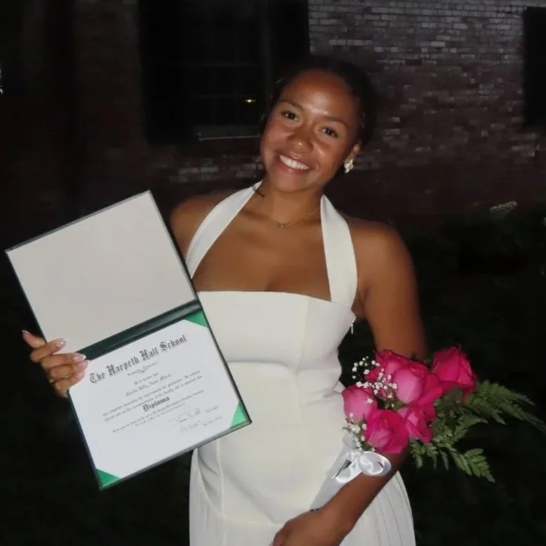 Young woman in white dress holding a diploma and a bouquet of pink roses, smiling at camera.