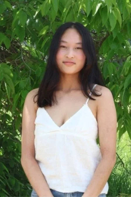 A young woman with long black hair, wearing a white spaghetti strap top, standing outdoors in front of green leafy trees.