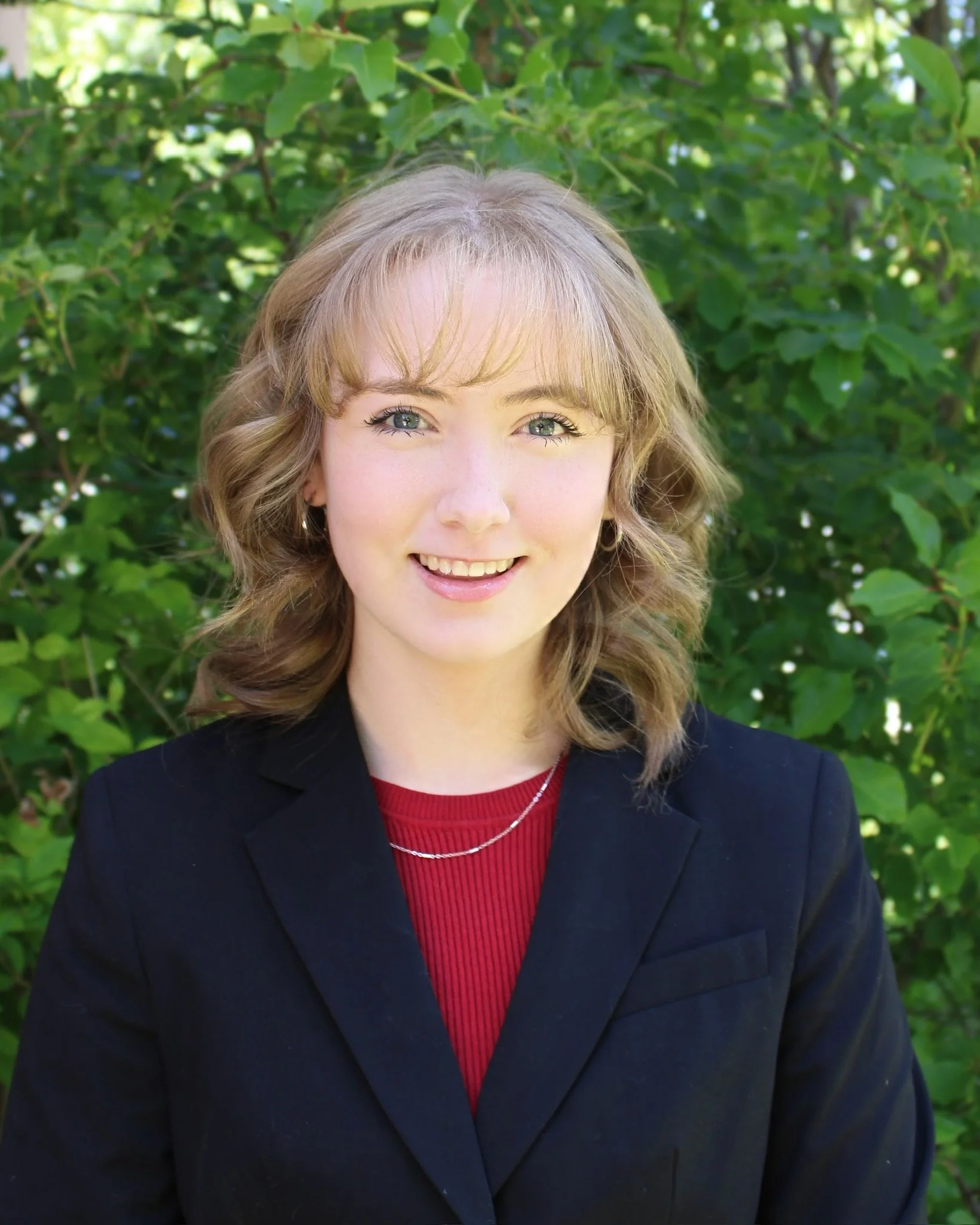 A woman with wavy blonde hair and green eyes smiling, wearing a black blazer and a red top, standing outdoors in front of green leaves and foliage.