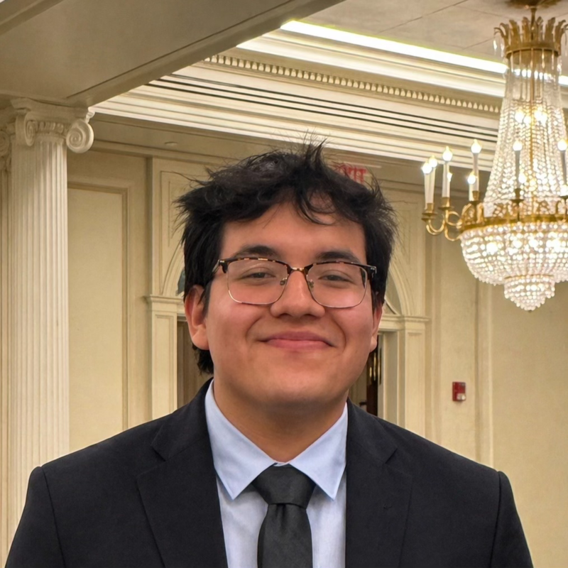 Young man wearing glasses, a suit, and a tie smiling at the camera in an elegant room with chandelier and ornate architectural details.