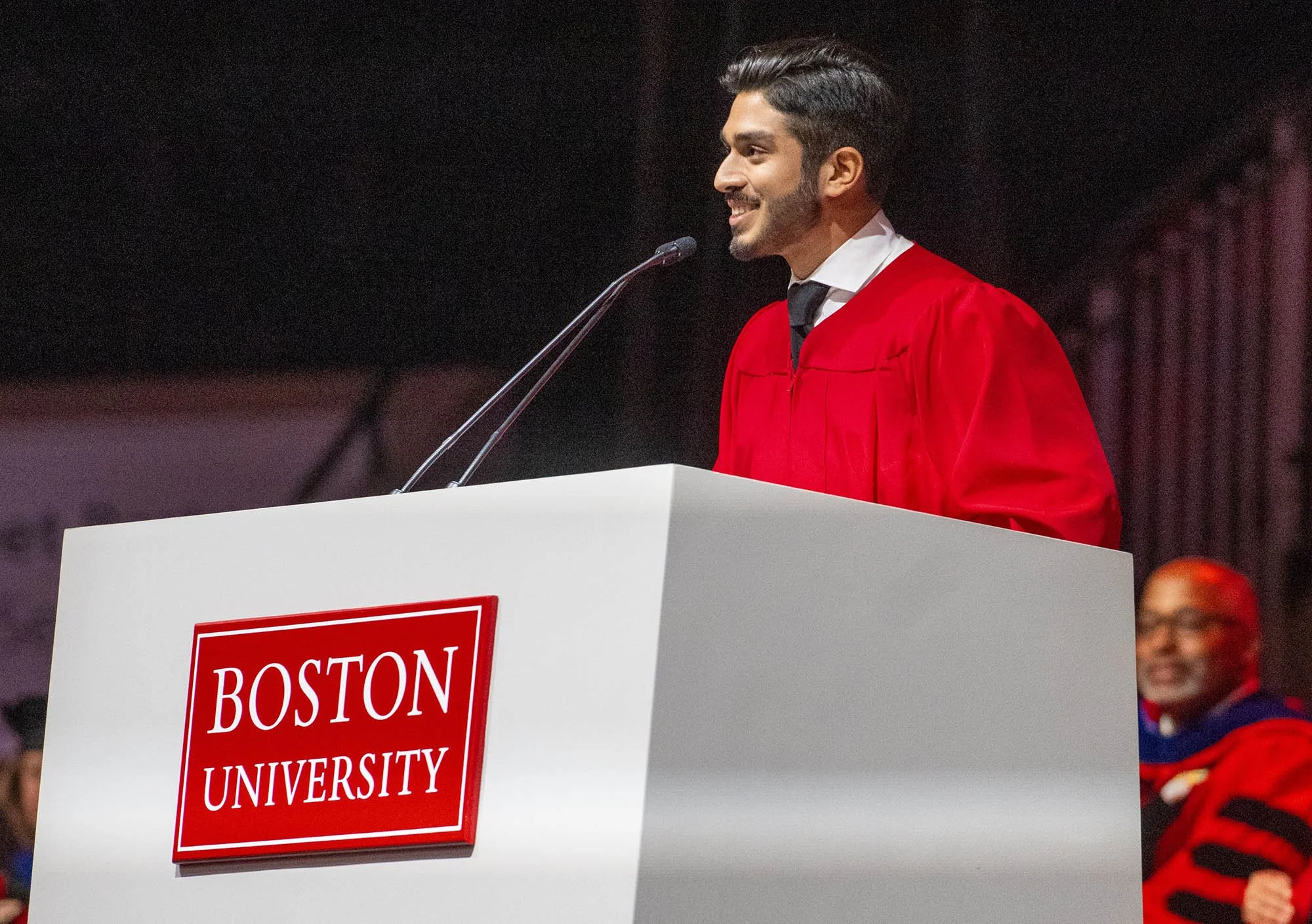 A young man with a beard and styled hair wearing a red graduation gown, speaking at a podium with a microphone at Boston University graduation ceremony.