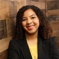 A young woman with curly hair smiling, wearing a black blazer and yellow top, standing against a wooden wall.