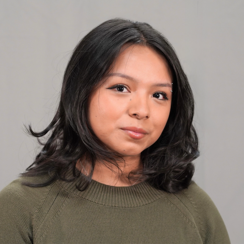 A young woman with shoulder-length black hair, wearing a green sweater, looking at the camera with a slight smile against a gray background.