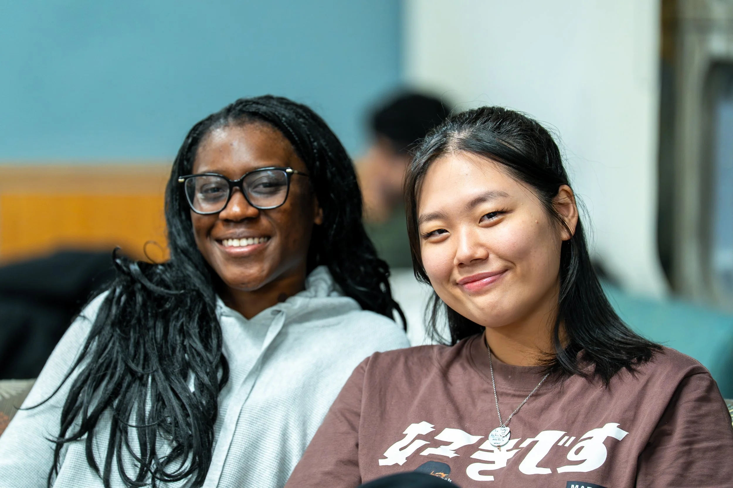 Two smiling young women, one of African descent with glasses and long black hair, and one of Asian descent with long black hair, posing closely together indoors.