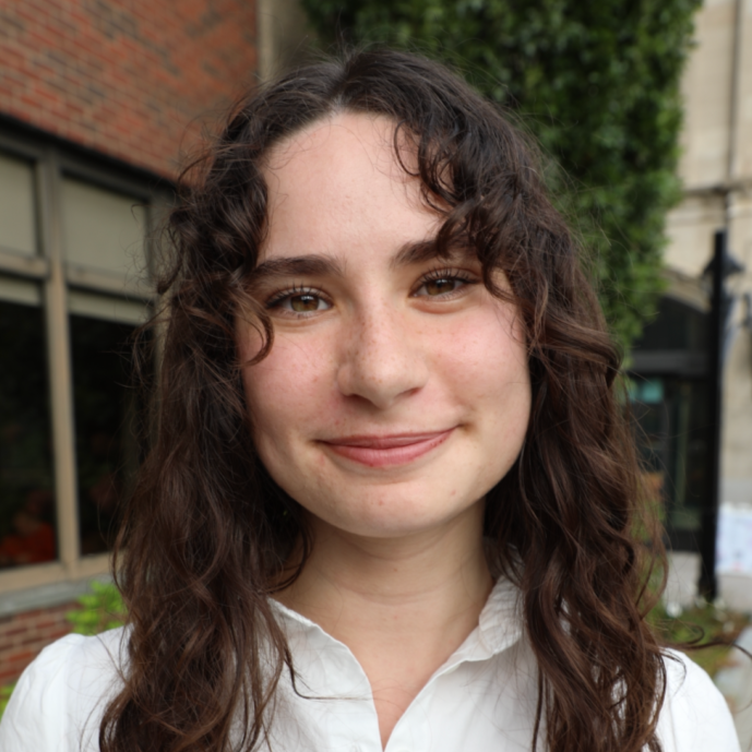 A young woman with long, curly brown hair smiling outdoors in front of a brick building and some green foliage.