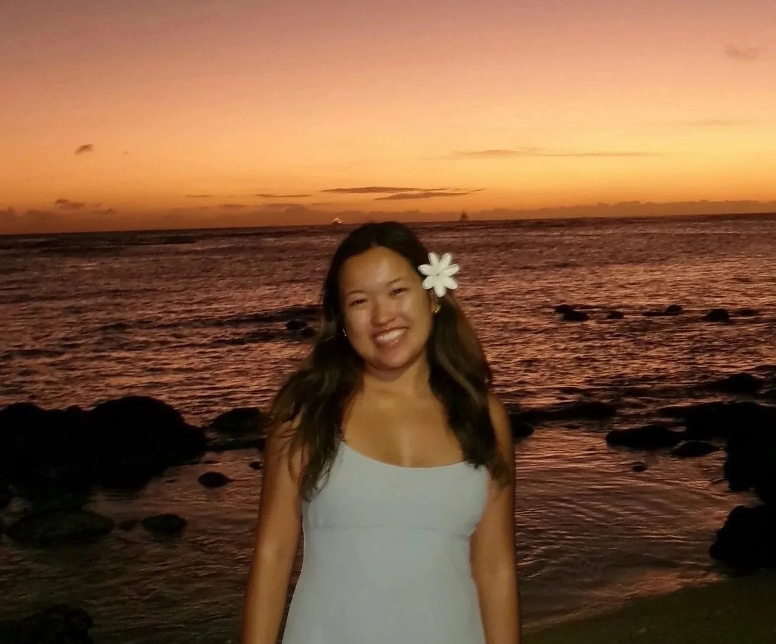 A woman with a white flower in her hair, smiling at the camera, standing by the ocean during sunset with a colorful sky and rocks in the background.