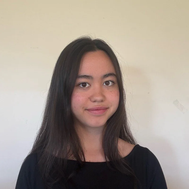 A young woman with long dark hair, wearing a black top, standing against a plain light-colored wall.