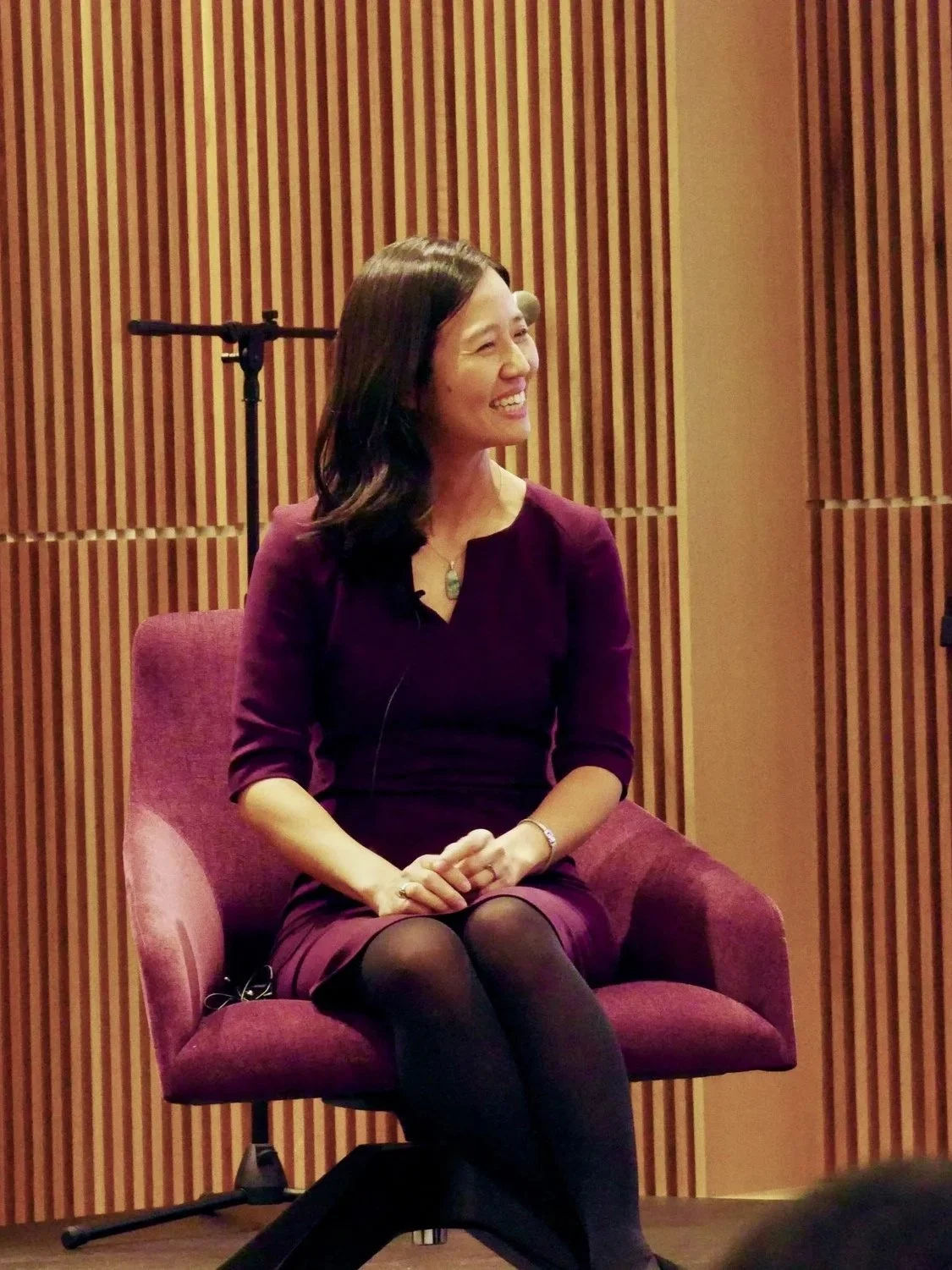 A woman with dark, wavy hair is sitting in a maroon chair, smiling, in front of a wooden backdrop with vertical slats.