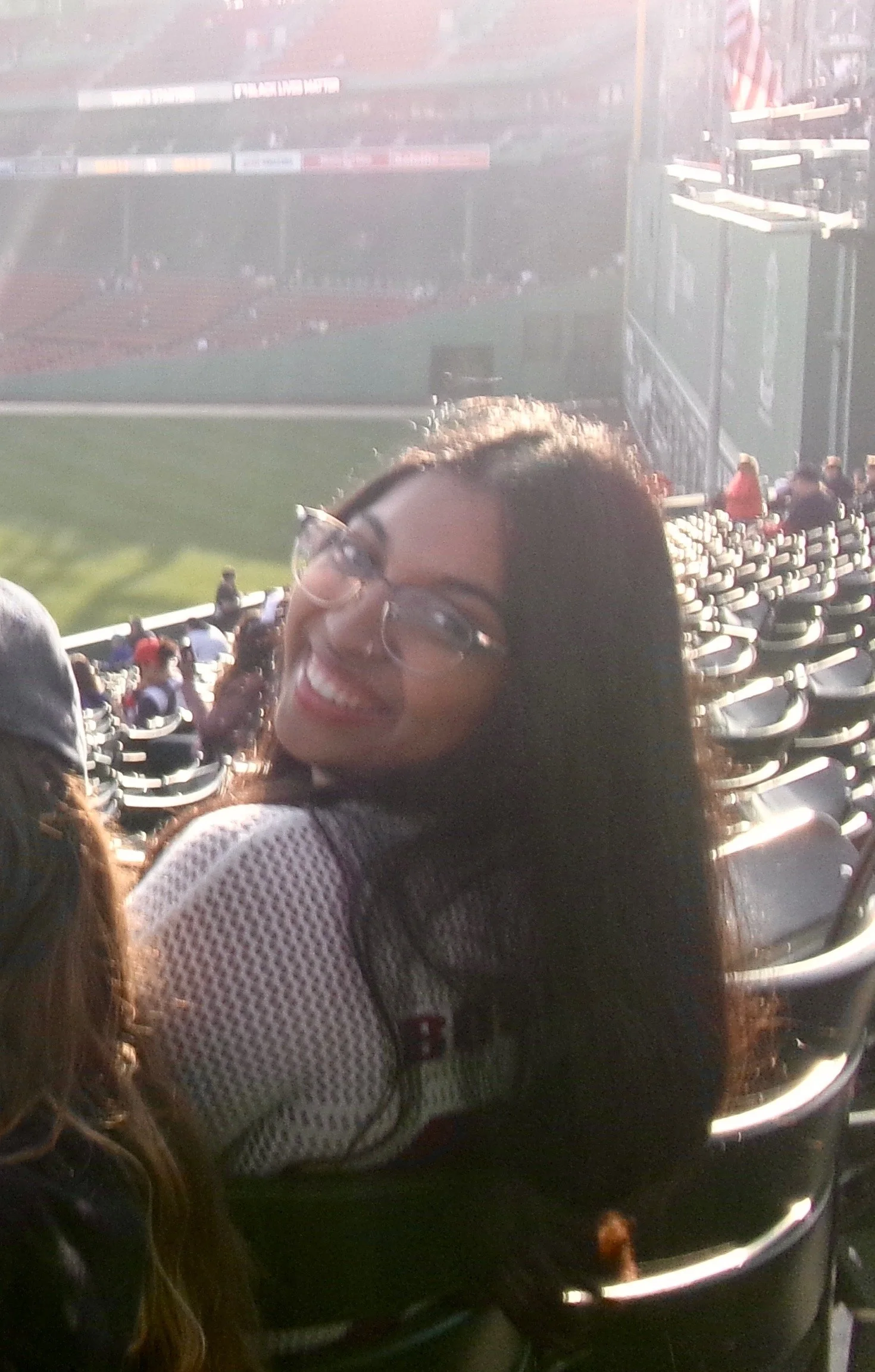 A smiling woman wearing glasses, sitting in a stadium with empty seats in the background, possibly during a sports event.