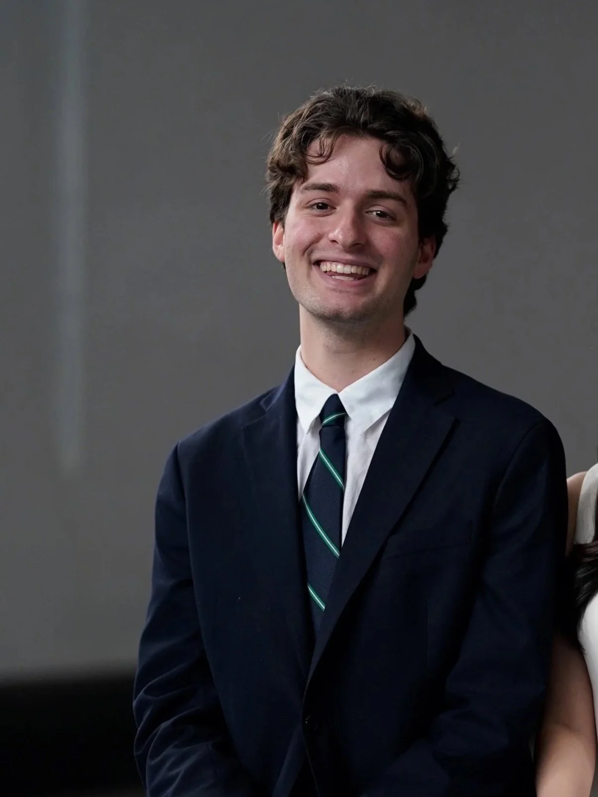 A young man in a dark suit and tie smiling at the camera against a plain gray background.
