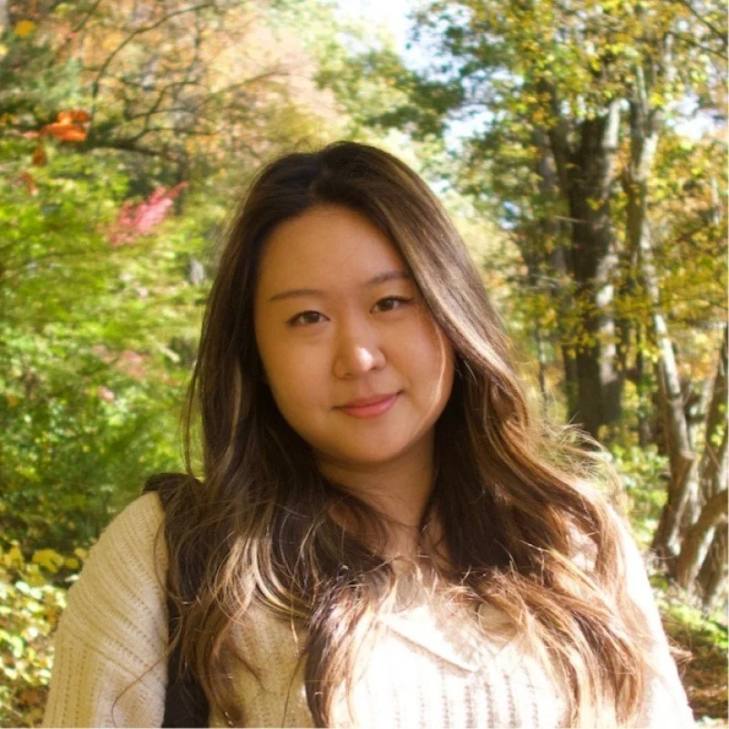 Young woman with long wavy hair standing outdoors among trees with autumn foliage.