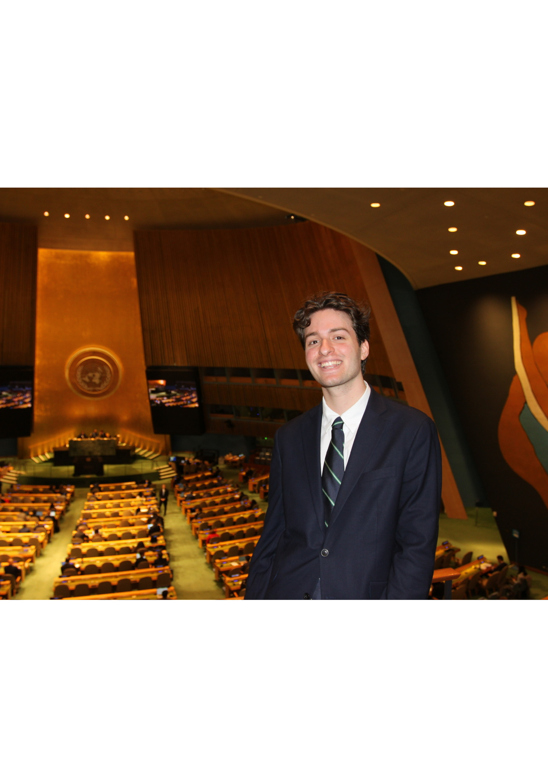 A young man in a dark suit, white shirt, and striped tie standing in a large conference hall with many empty seats, a large circular emblem on a wooden wall, and a decorated stage in the background.